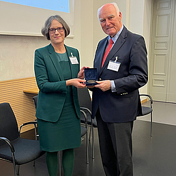 Michael Hoffmann-Becking trägt einen dunkelfarbenen Anzug, eine rot-weiß-gemusterte Krawatte und ein hellblaues Hemd. Neben ihm steht Julia Blumenthal, die ein grünes Kostüm trägt. Sie überreicht ihm die Humboldt-Universitäts-Medaille.